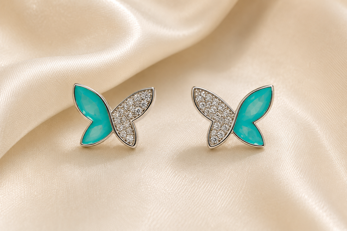 A pair of butterfly-shaped stud earrings with bright turquoise blue enamel wings and silver pavé-set back wings, displayed on a draped cream satin fabric.