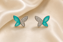 A pair of butterfly-shaped stud earrings with bright turquoise blue enamel wings and silver pavé-set back wings, displayed on a draped cream satin fabric.